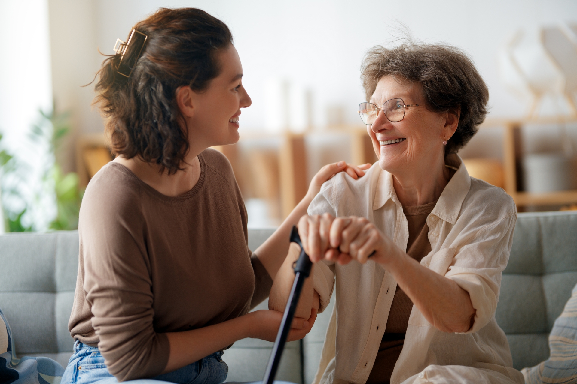 Older woman with a cane talking with a younger woman on a sofa.